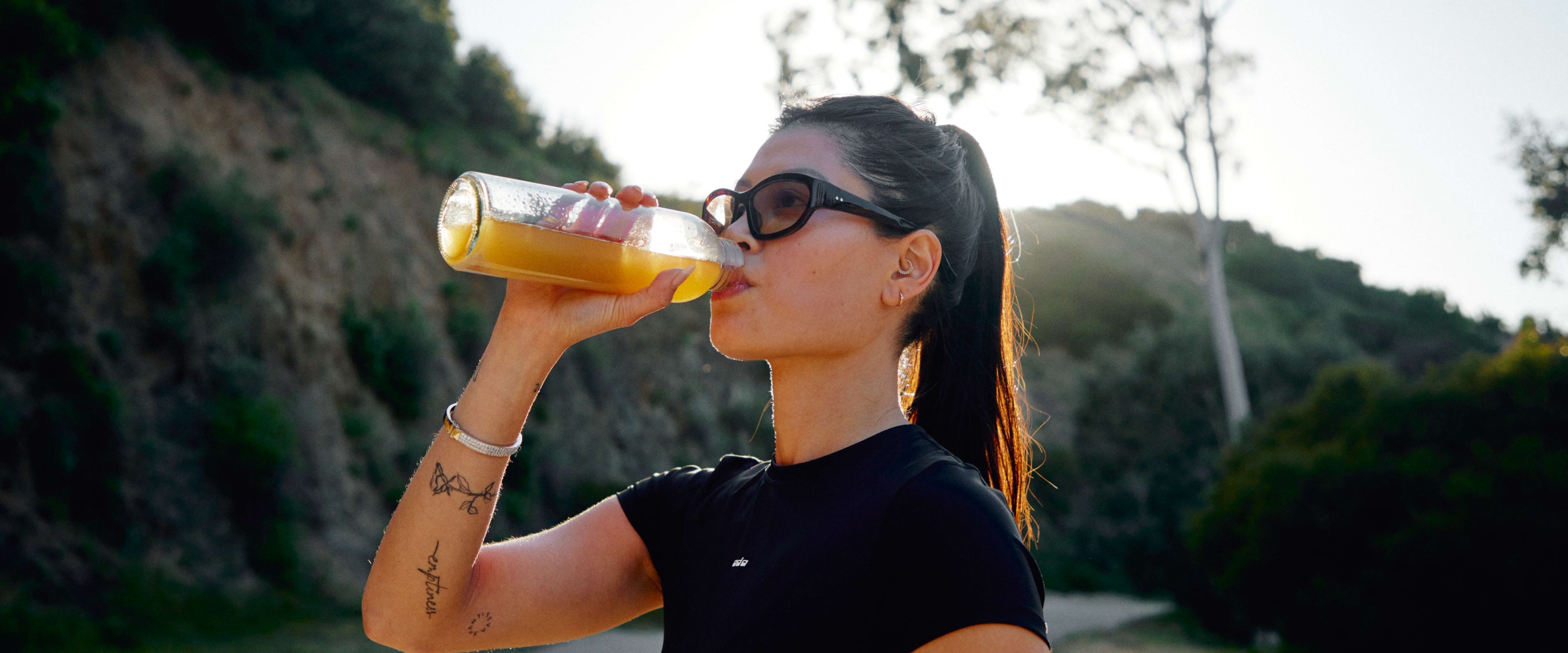 Close up photo of athletic woman with sunglasses drinking Moxie from a clear glass bottle