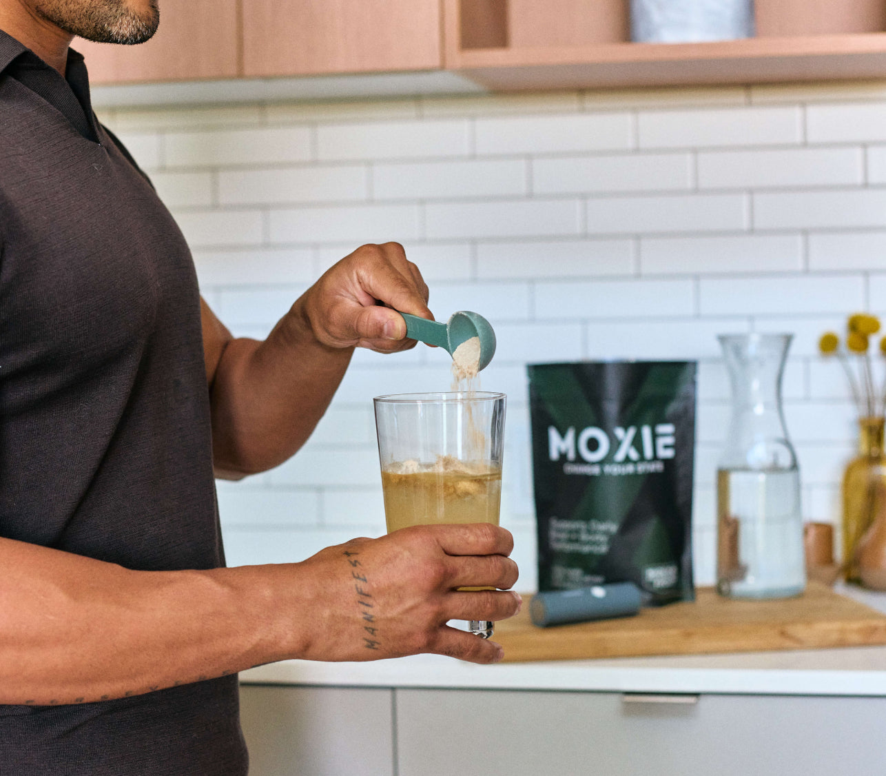 Close-up photo of man pouring Moxie powder into a glass of water in a kitchen with Moxie packaging on counter in the background